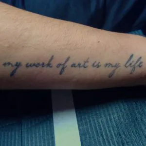 A photo of a script tattoo on the forearm, reading "My Work of Art is My LIfe" before any treatments.